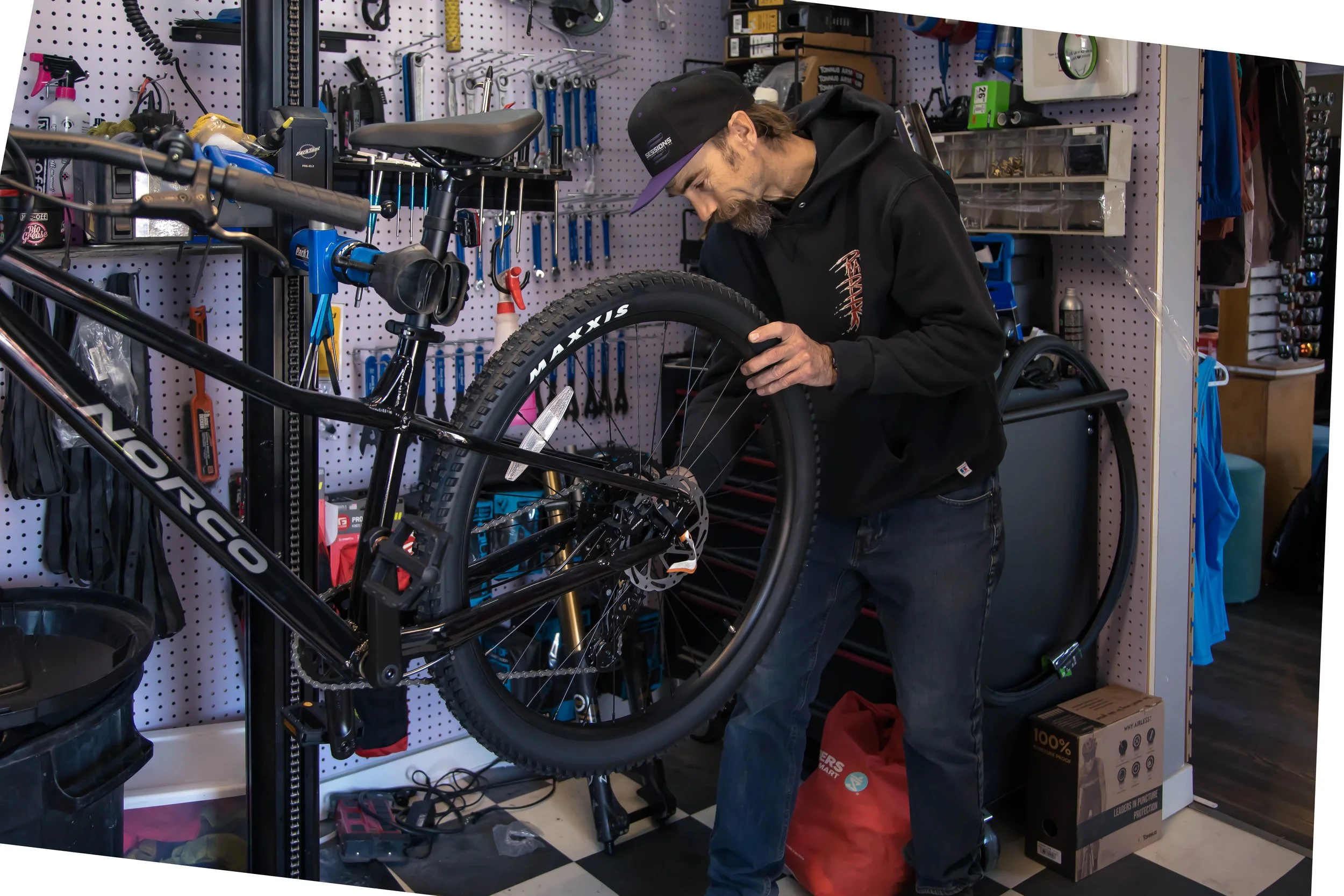 A man working on repairing a mountain bike in a workshop surrounded by tools, with a pegboard wall, shelving, and a red bag on the floor.