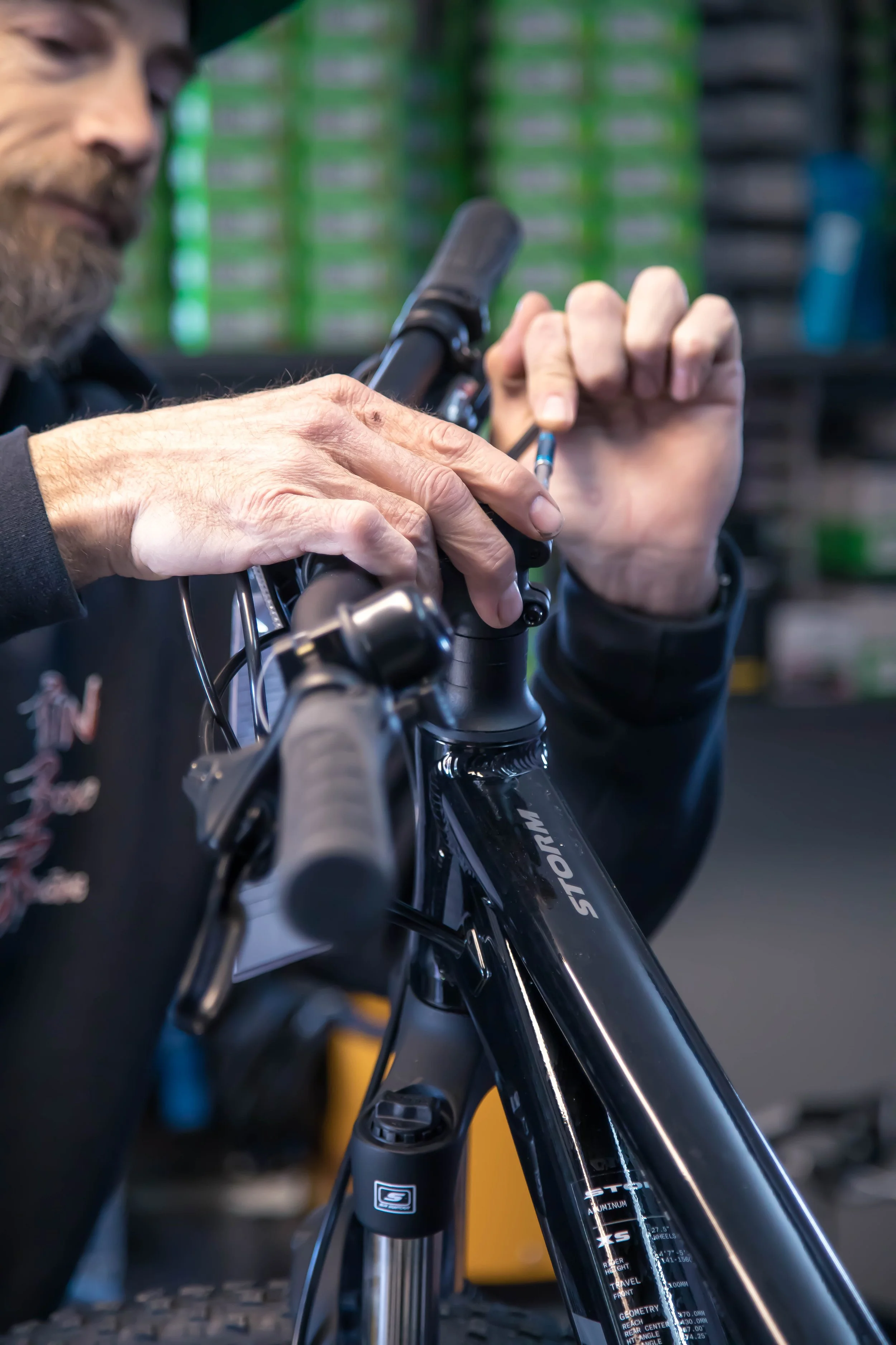 Close-up of a man working on a black mountain bike in a bike shop, adjusting the handlebar with shelves of tools and equipment in the background.