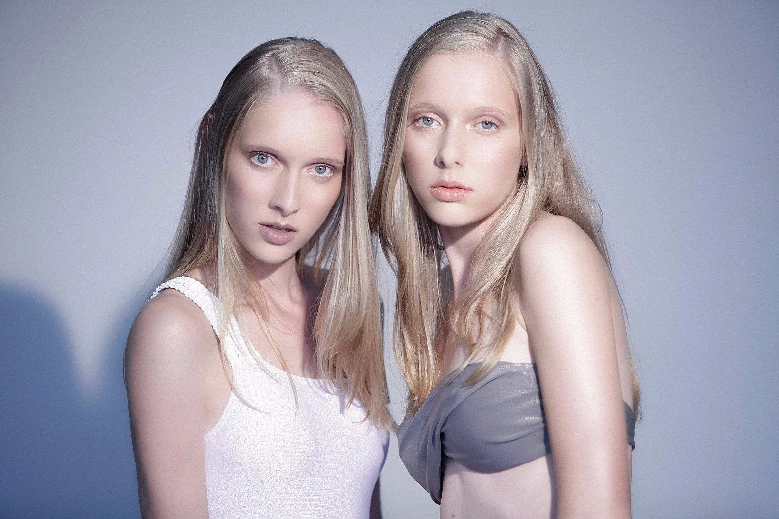 Two young women with blonde hair and fair skin posing against a plain gray background.