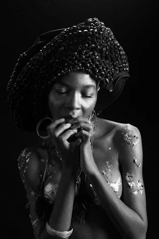 A woman with elaborate braided hair and glitter on her shoulder, looking down with her hand near her face, in black and white.