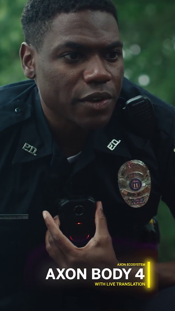 Close-up of a police officer in uniform with a badge, holding a handheld device, outdoors with blurred greenery in the background, caption reads 'AXON BODY 4 with live translation'.