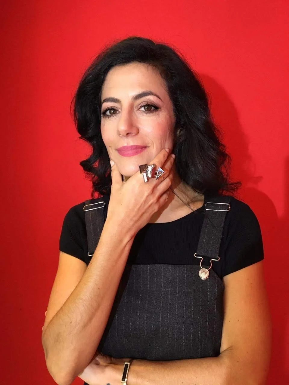 A woman with black hair and fair skin, wearing a black shirt and dark overalls, poses with her hand on her chin against a red background. She has rings on her fingers and a bracelet on her wrist, and smiles softly.