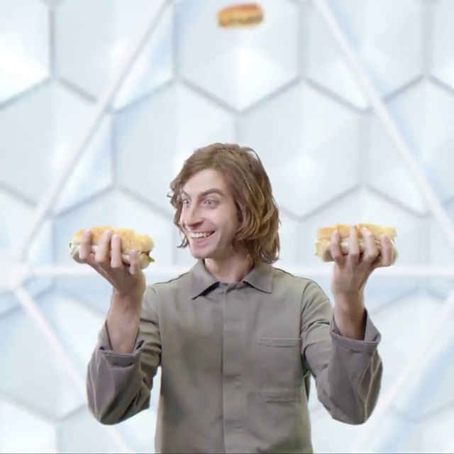 A man with long, wavy hair smiling and holding two sandwiches, one in each hand, in front of a geometric white background.