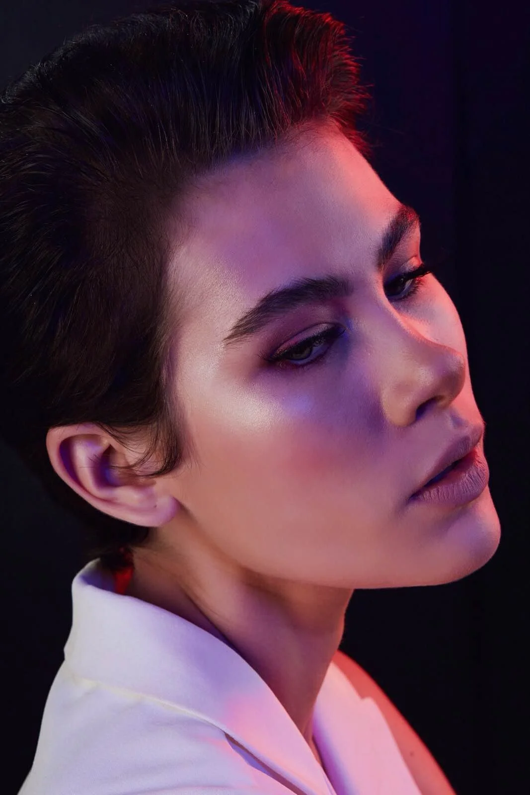 Close-up of a young woman with short, dark hair, wearing a white top, with soft, colorful lighting on her face against a dark background.