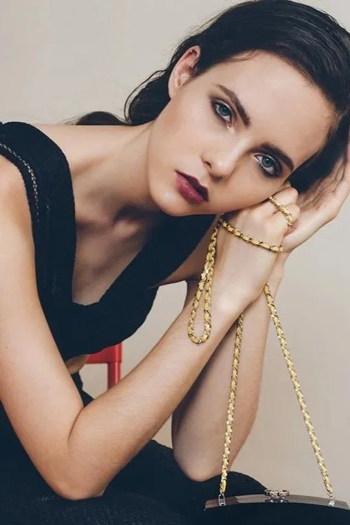 A young woman with dark hair and blue eyes, wearing a black sleeveless top, gold jewelry, and holding a gold chain purse, poses with her head resting on her hand.
