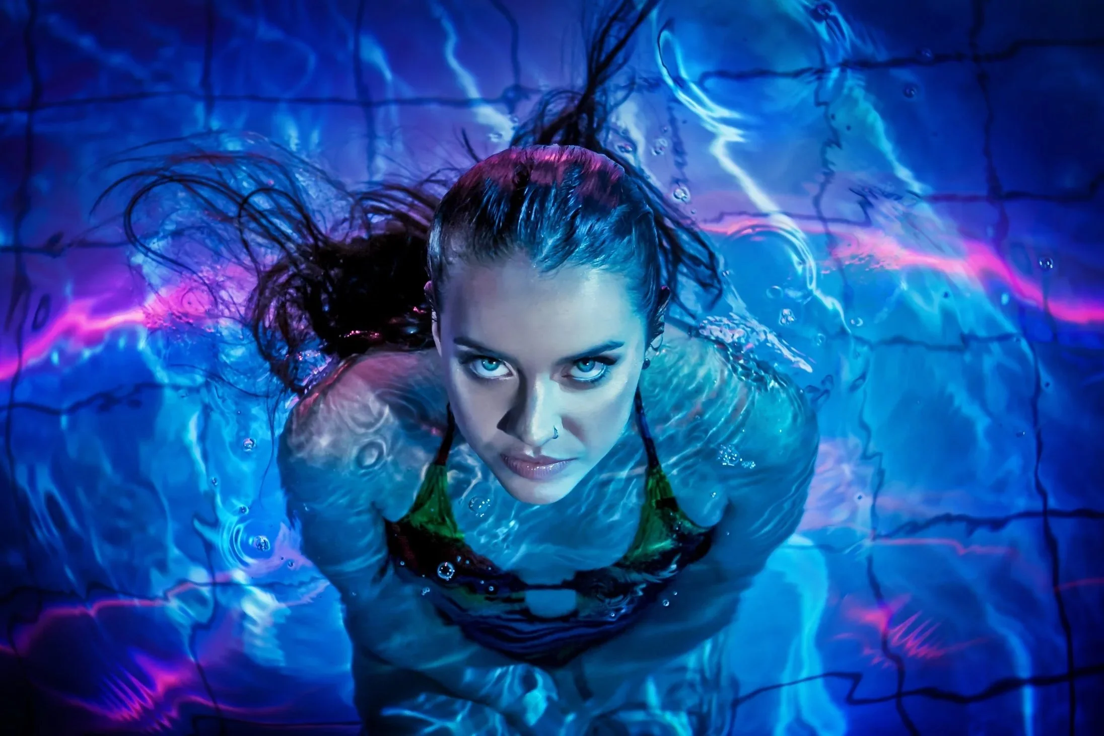 A woman with blue eyes and wet hair swimming in a pool with colorful neon lighting.