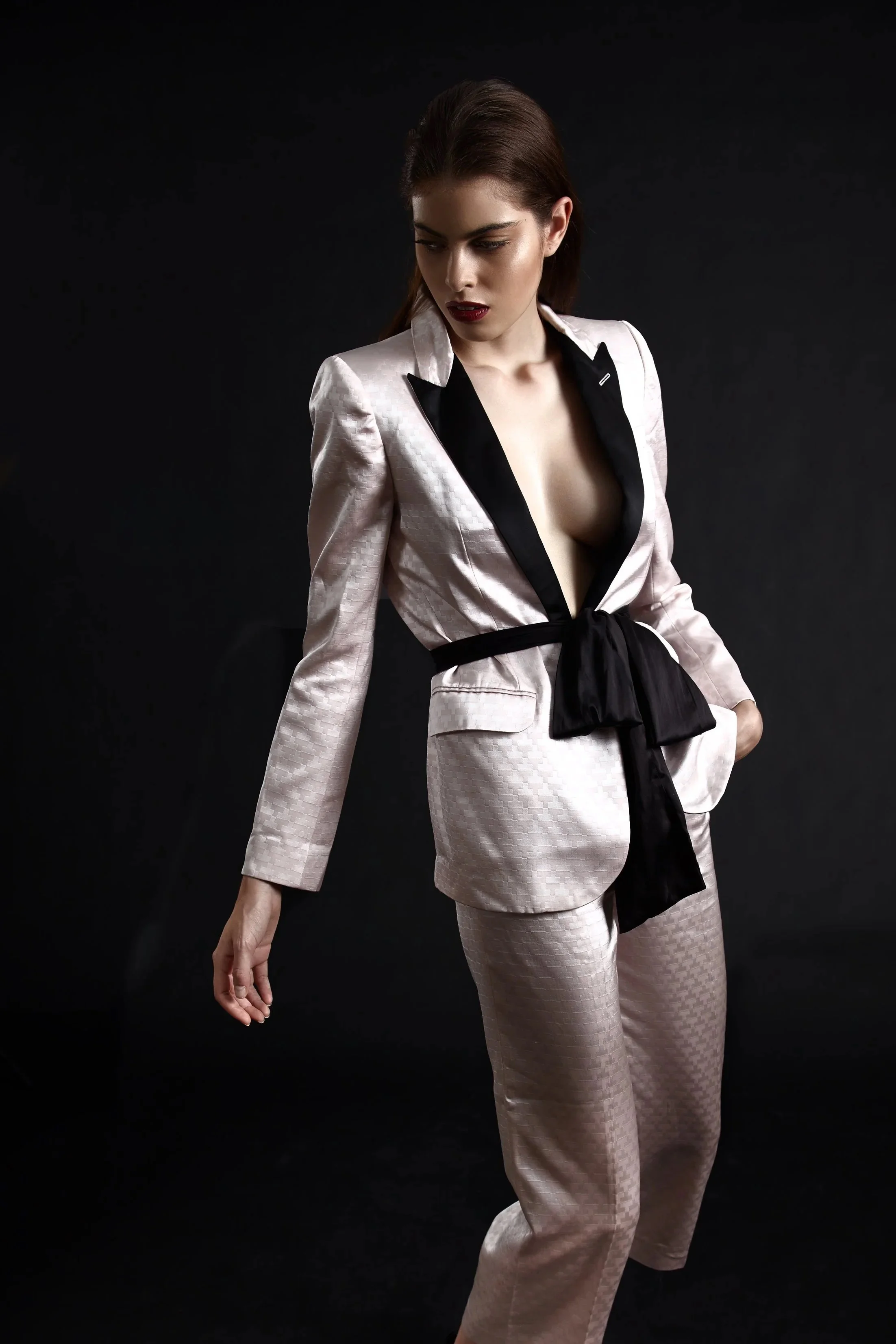 A woman in a light-colored satin suit with a black satin belt and lapels, and a revealing black inner shirt, standing against a black background.