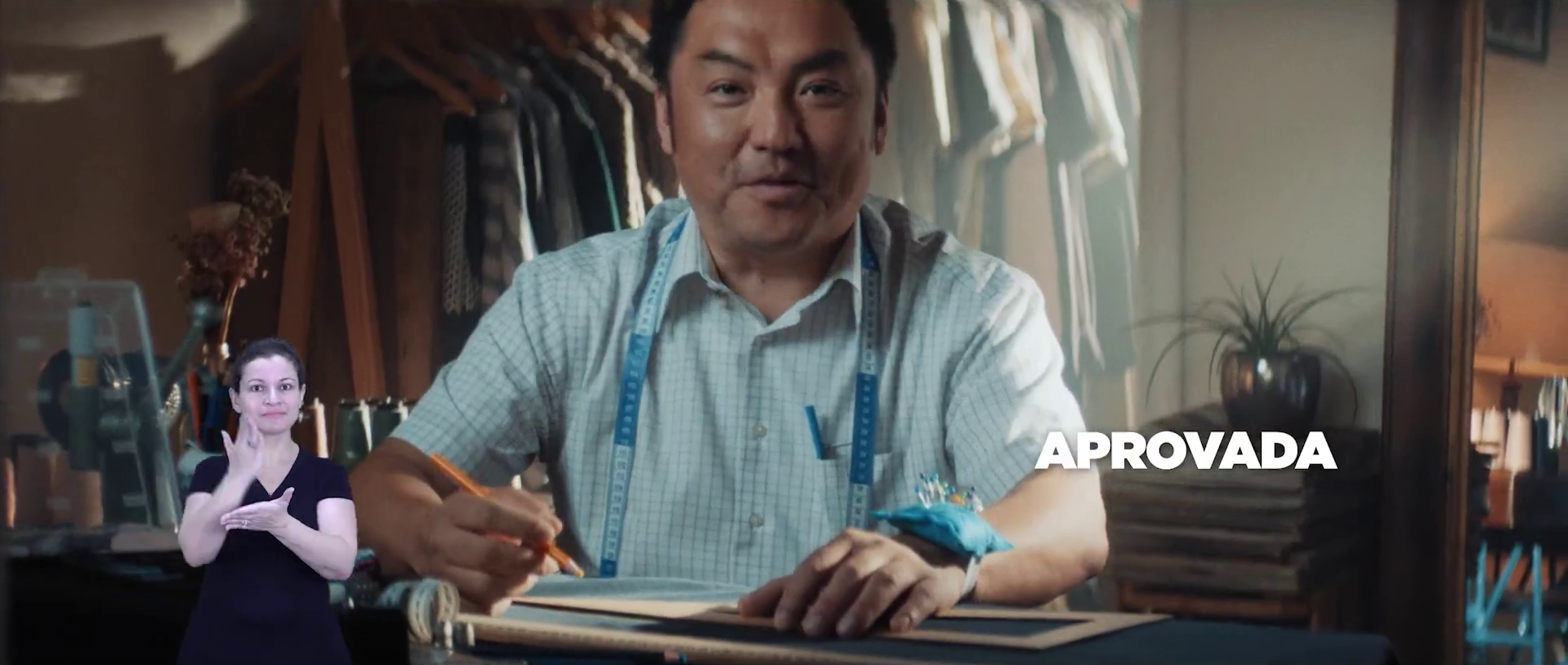 A man sitting at a desk with a tape measure around his neck, writing on a notebook, in a room filled with clothes, plants, and a lamp. The word 'APROVADA' is overlaid on the image.