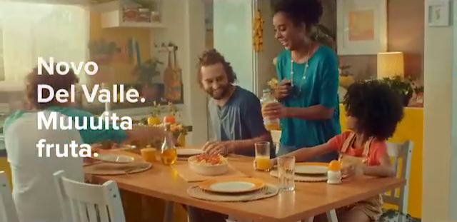 A family gathered around a dining table enjoying breakfast, with a woman serving juice, man smiling, and a young girl reaching for food, in a bright kitchen.