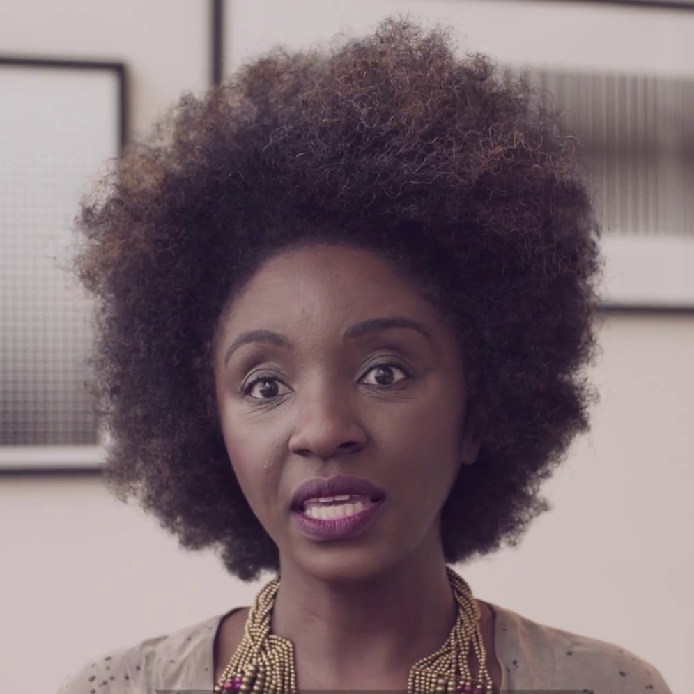 A woman with a large curly afro hairstyle, wearing a beaded necklace and a beige top, speaking in front of framed pictures on a wall.
