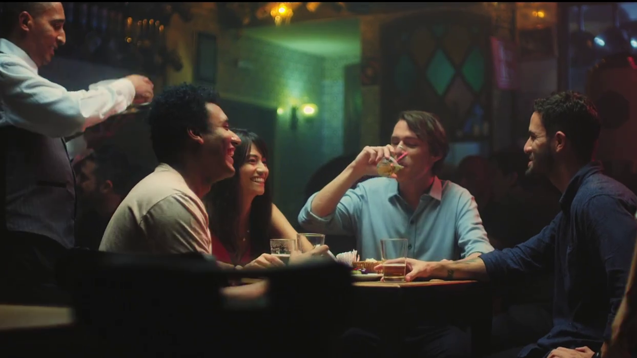 Four friends enjoying drinks and laughing together at a bar or restaurant, with a waiter serving drinks in the background.