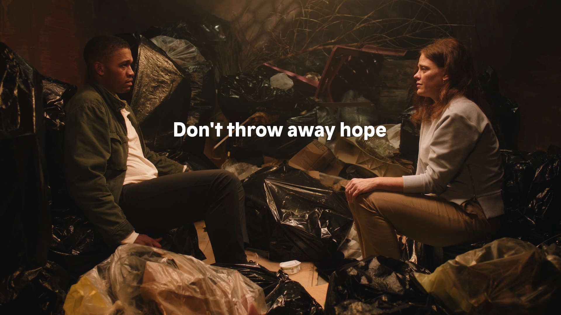 A woman and a young man sit facing each other in a dimly lit room filled with garbage bags. They appear to be having a serious conversation, with the woman speaking and the young man listening. The scene is intense and emotional, with the text overla