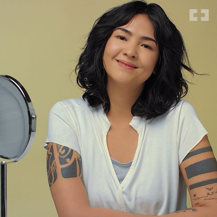A woman with shoulder-length dark curly hair smiling and looking at the camera, wearing a white shirt with rolled-up sleeves, sitting in front of a small mirror, with a yellow-green background.