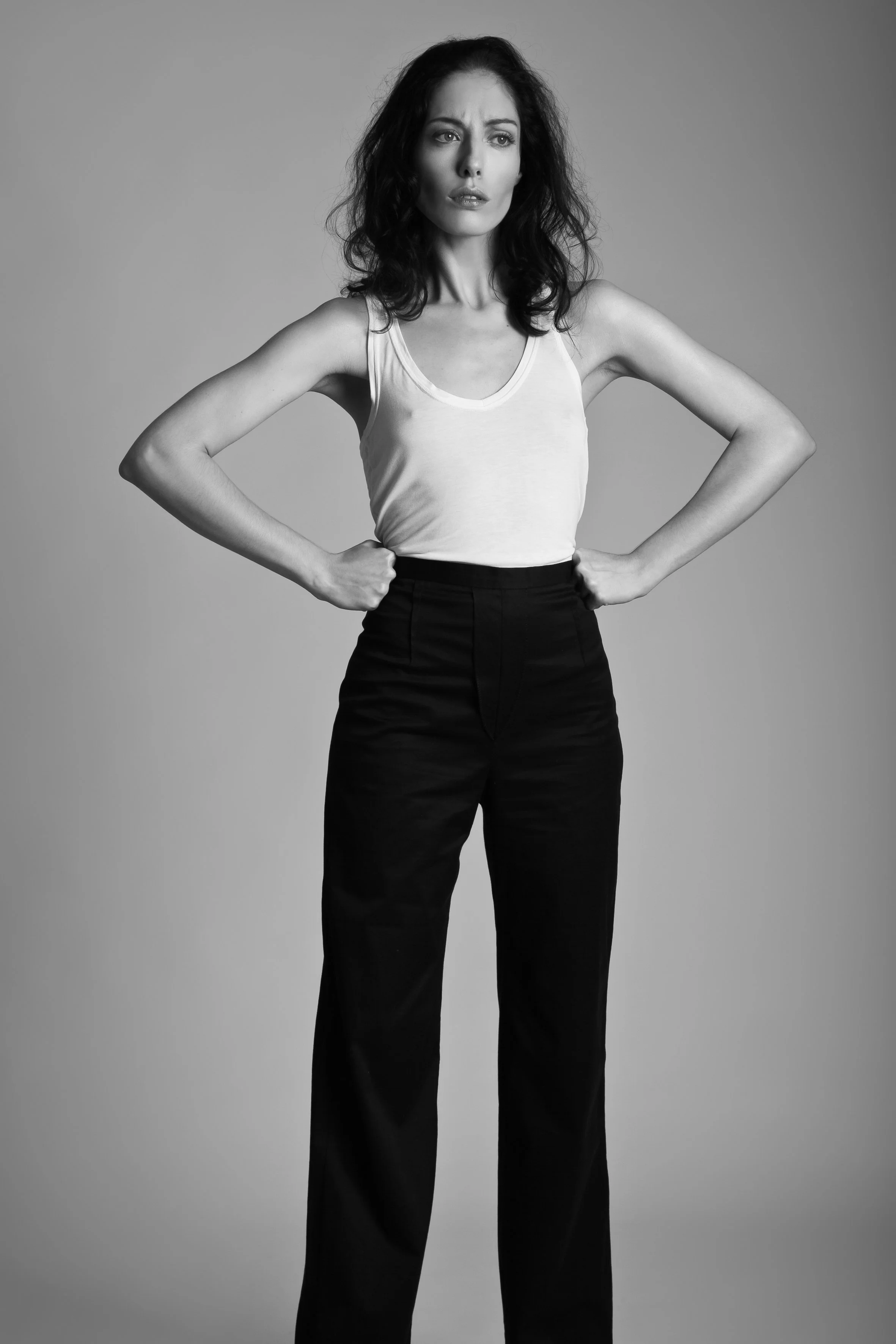 Black and white photo of a woman standing with hands on hips, wearing a sleeveless top and high-waisted pants, with a serious expression.