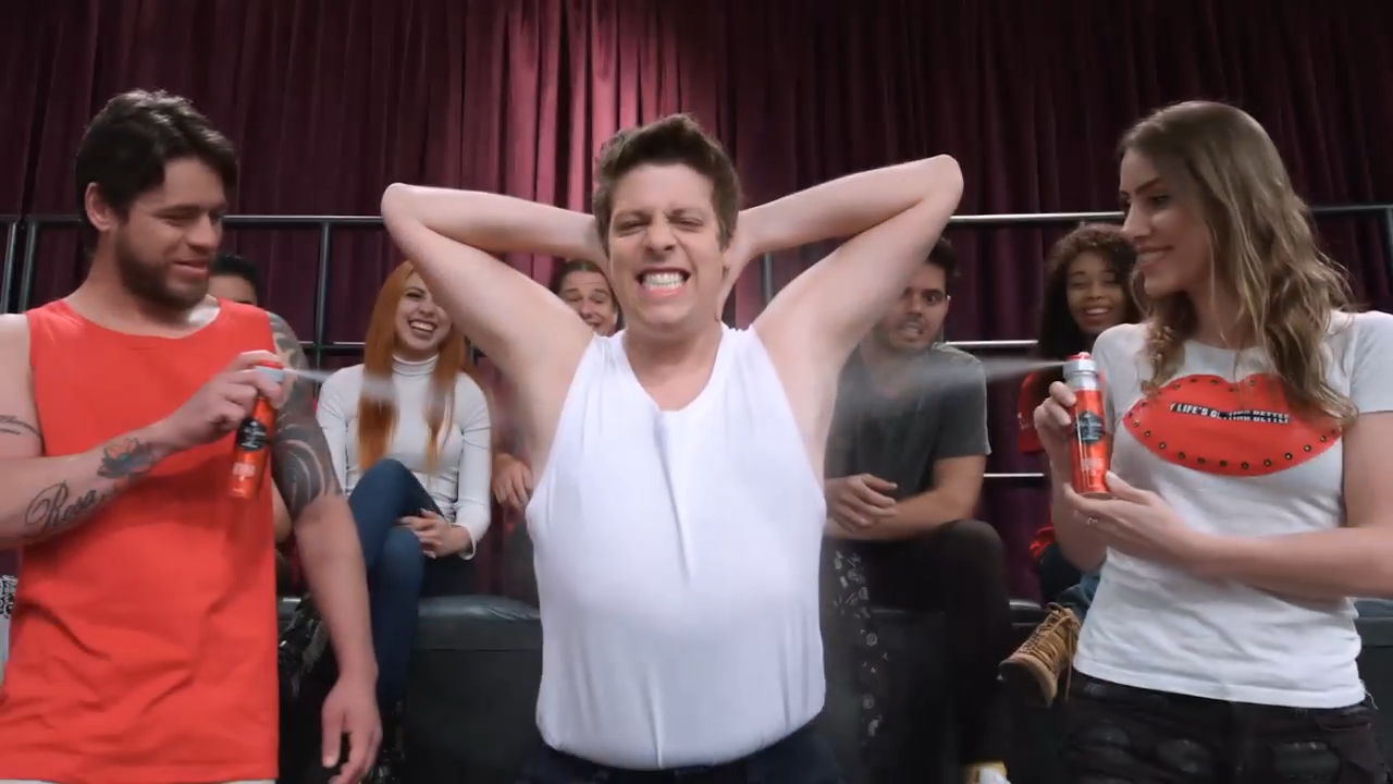 A group of people laughing and enjoying themselves during a wet, fun activity. One man in a white tank top is in the center, with his hands behind his head, squinting and smiling despite being wet. Two women, one on each side, hold spray bottles, and