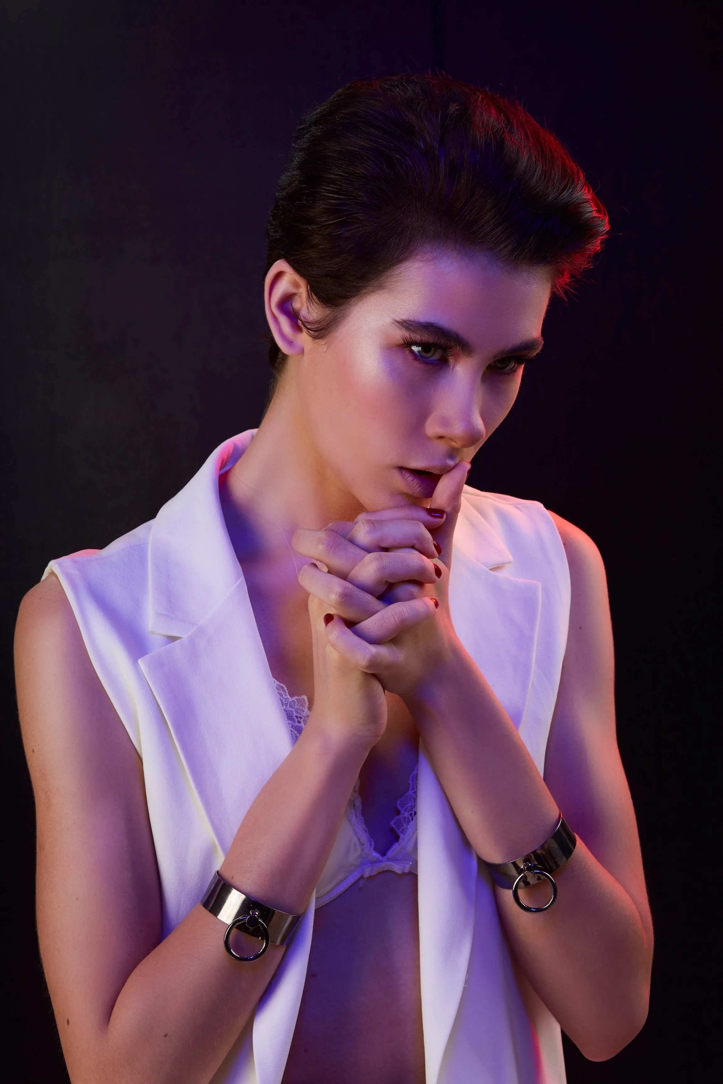 A woman with short dark hair and striking eyes is posing with her finger on her lips. She is wearing a white sleeveless blazer and black metal wrist cuffs with rings, standing against a dark background with colorful lighting.
