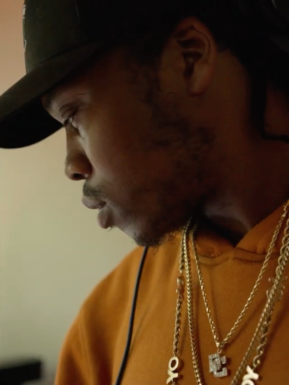 Close-up side profile of a young man wearing a black cap, gold chains, and an orange hoodie, looking down with a neutral expression.