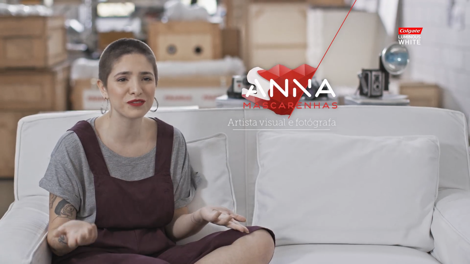A woman with short hair and tattoos on her arms sitting on a white couch, speaking, with a background of boxes and studio equipment. There is overlay text with a red logo and the words "ANA MASCARENHAS Artista visual e fotógrafa" and a small logo for