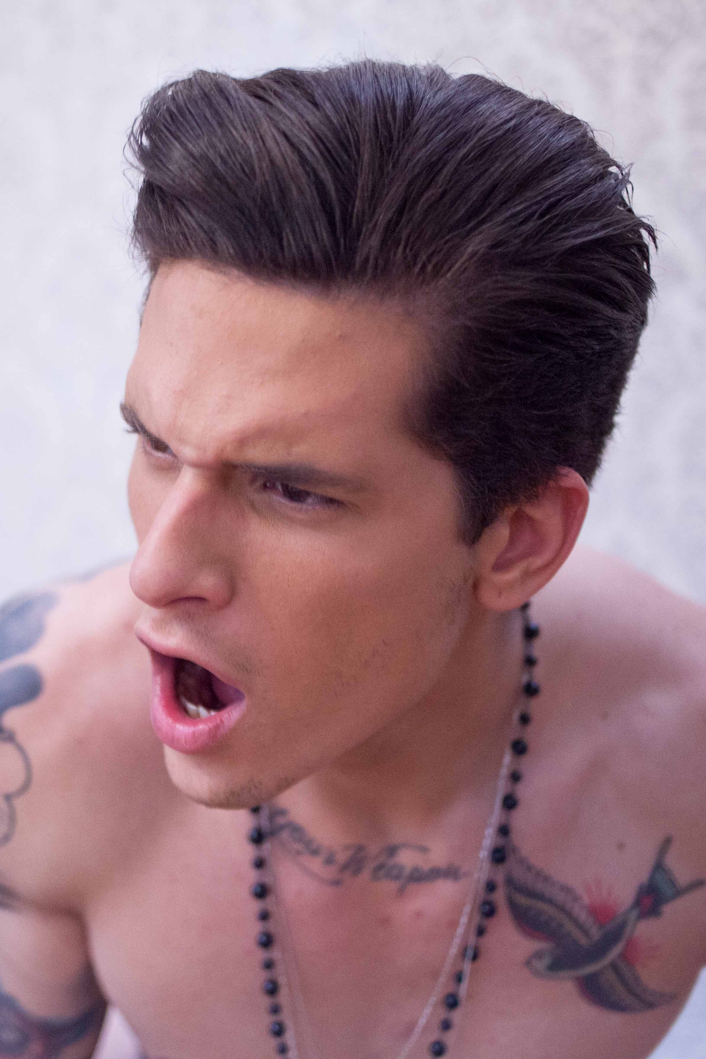 A shirtless man with ink tattoos on his shoulders and chest, wearing a beaded necklace, has dark hair slicked back and is yelling or shouting with an intense facial expression.
