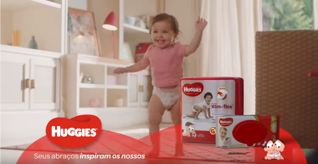 A happy toddler in a pink shirt and underwear playing with Huggies diapers on a red table in a well-lit living room.