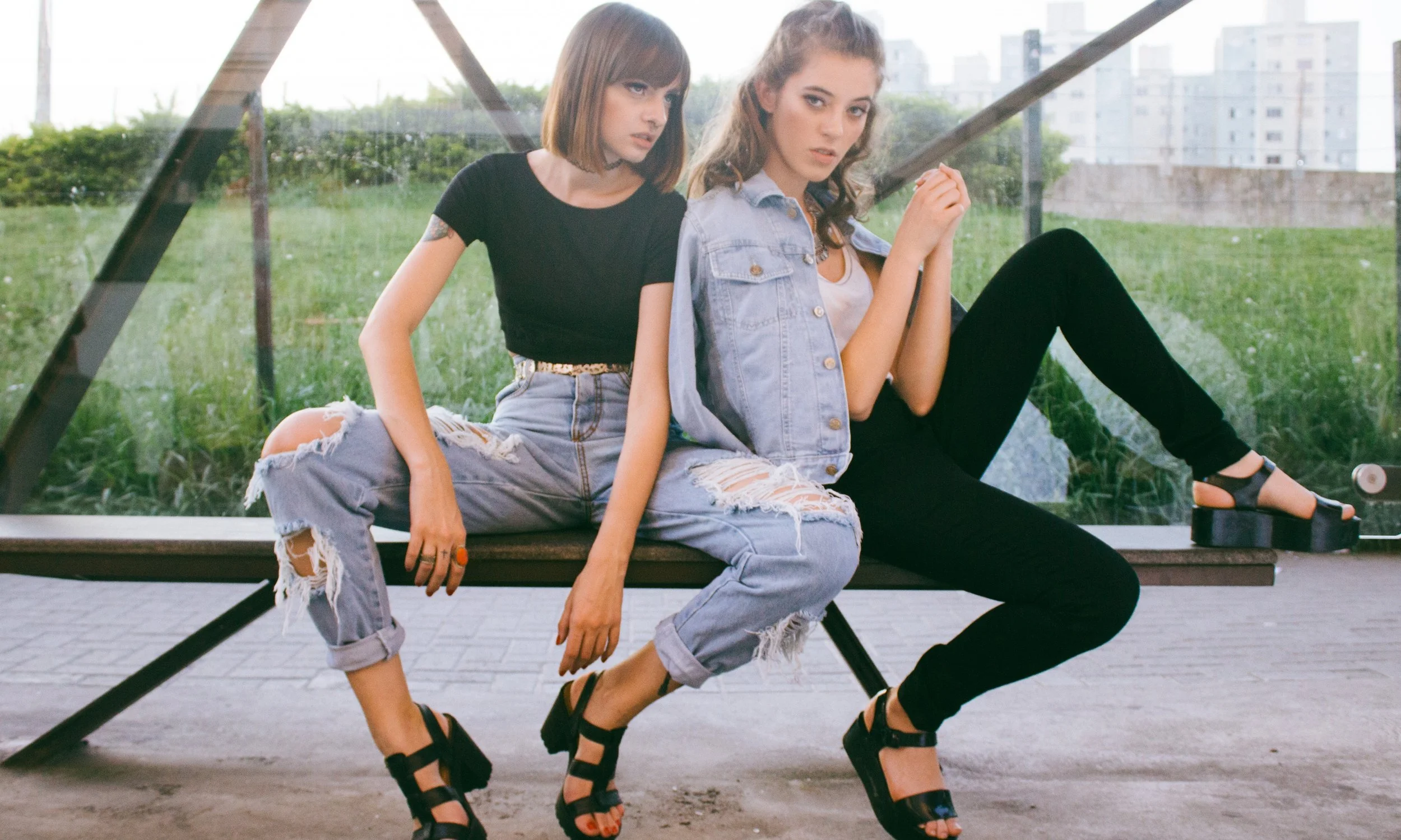 Two young women sitting on a bench in front of a glass window with greenery and buildings outside. One is dressed in a black crop top and distressed jeans, the other in a denim vest and black pants, both wearing black platform sandals.