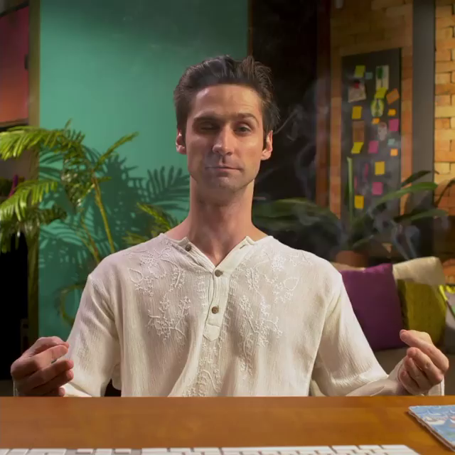 A man with dark hair is sitting at a table, making a gesture with his hands in a calm indoor setting with plants and colorful decor in the background.