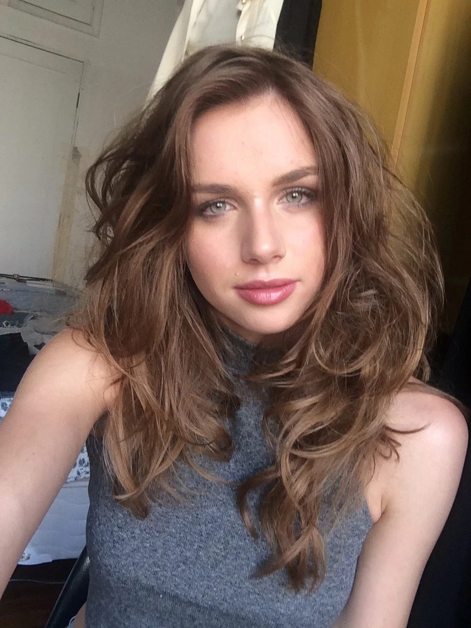 A young woman with long, wavy brown hair and blue eyes taking a selfie indoors, wearing a sleeveless gray top.