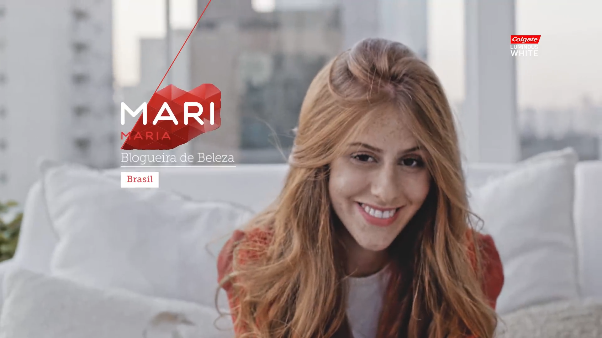 A smiling woman with long red hair sitting on a white couch in a bright room with large windows.