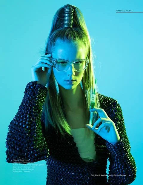 Fashion model with long hair in a high ponytail, wearing glasses and a shiny black jacket, holding a test tube.