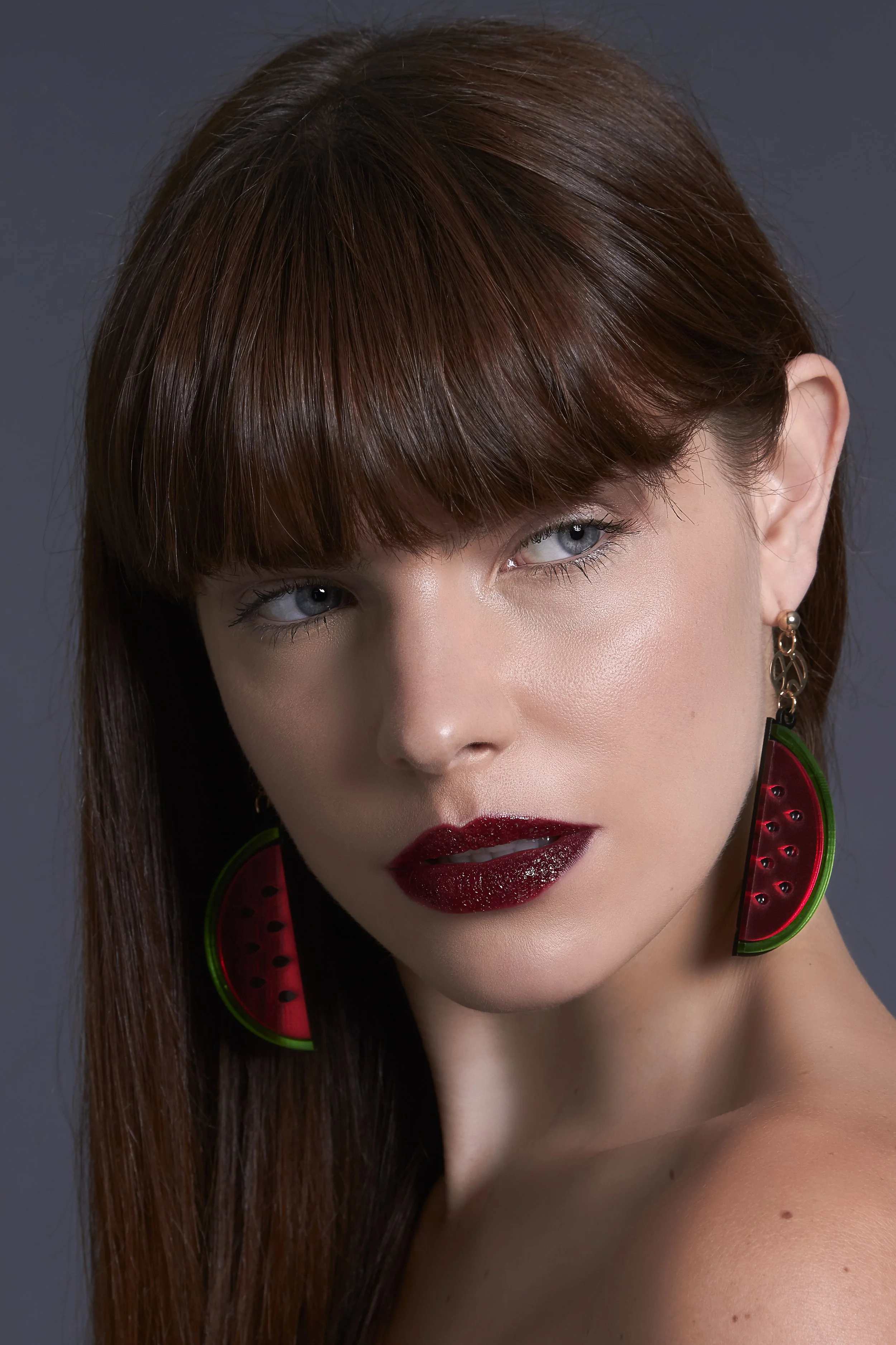 A woman with straight auburn hair and blue eyes wearing watermelon-shaped earrings and dark red lipstick.