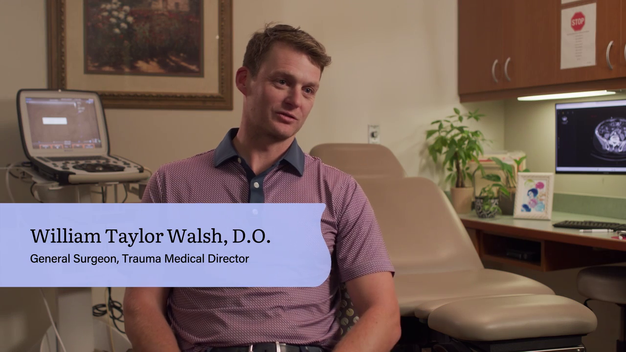 A man sitting in a medical examination room with a computer monitor displaying a scan, and medical equipment on a cart behind him. Text overlay identifies him as William Taylor Walsh, D.O., General Surgeon and Trauma Medical Director.