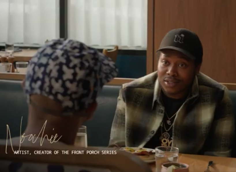 Two men having a conversation at a restaurant table, one wearing a baseball cap and plaid shirt, the other with patterned headwear, with food and drinks on the table.