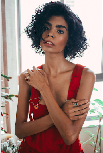 Woman with curly black hair wearing a red sleeveless top, standing near a window with green plants in the background.