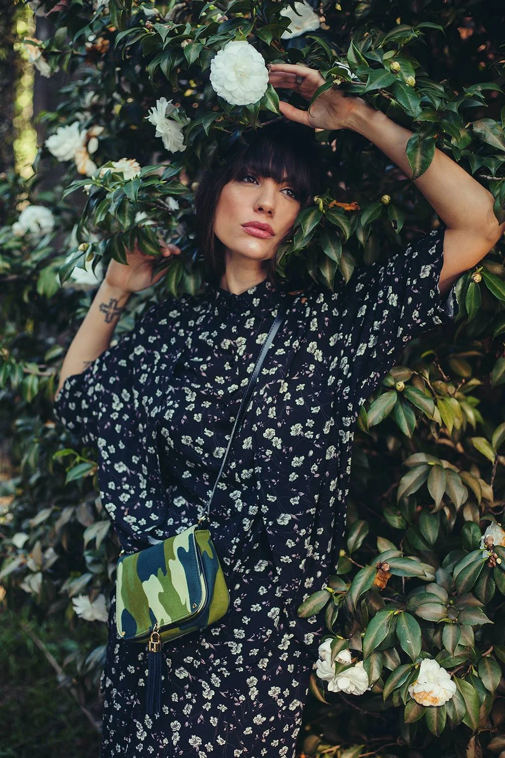 A woman with dark hair and bangs, wearing a black dress with white floral patterns, stands among green bushes with large white flowers, holding a branch above her head.
