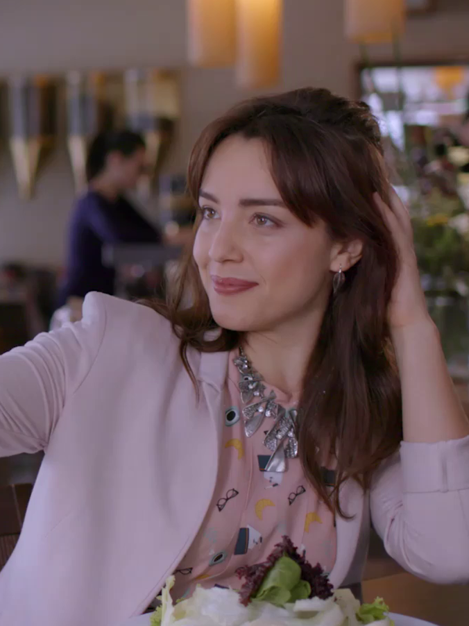 Woman with brown hair sitting at a table in a restaurant or cafe, smiling and touching her hair, with a backdrop of blurred interior and another person in the background.