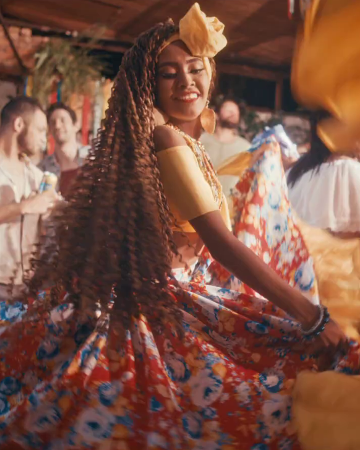 A woman with long, curly hair dancing at a festive party, wearing a bright yellow top and floral skirt, with people in the background.