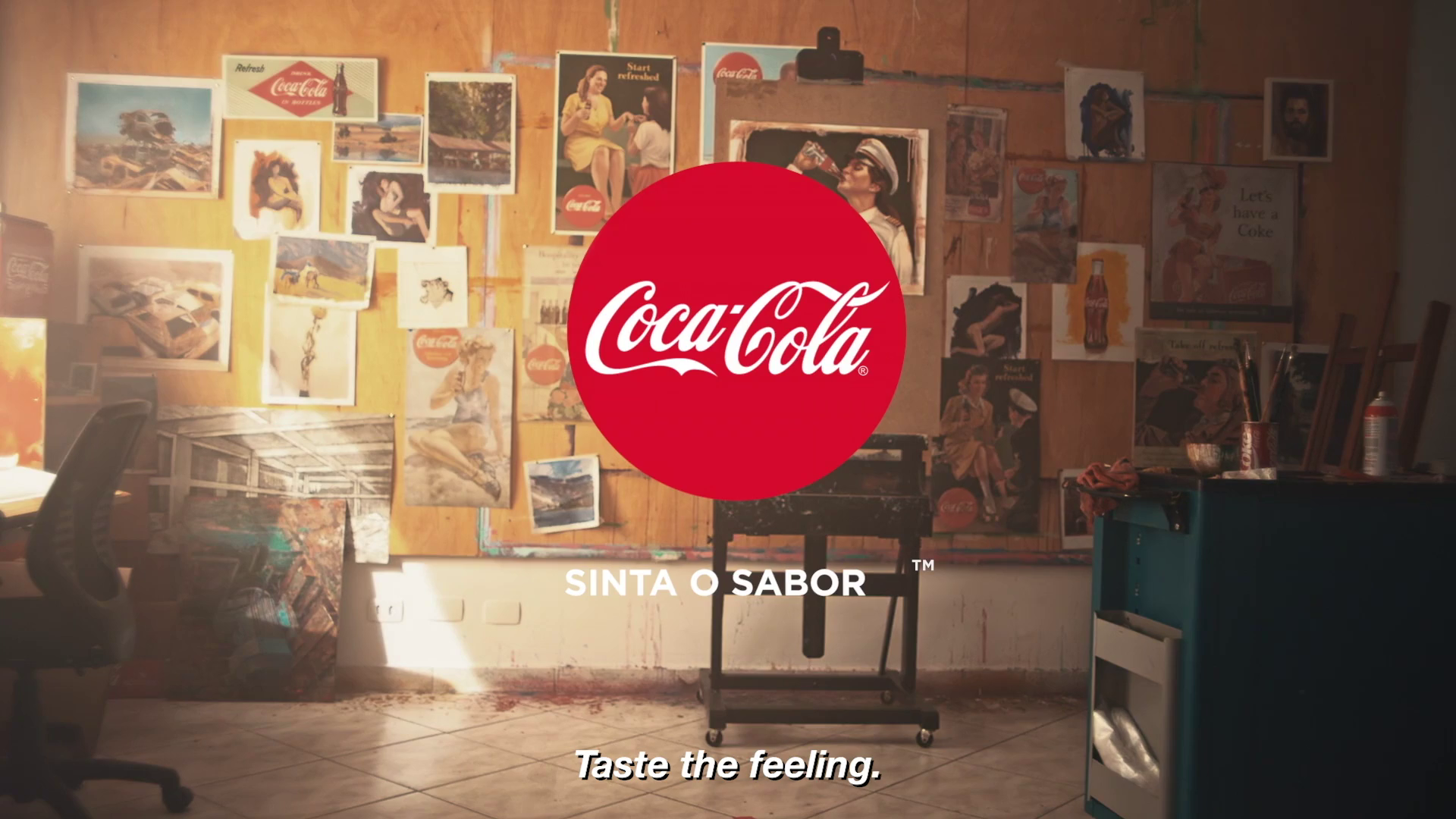 A room with a corkboard wall covered in various vintage posters and pictures. There are chairs, a desk, and art supplies on the right side. A large Coca-Cola logo is centered, with the slogan 'Taste the feeling' at the bottom.