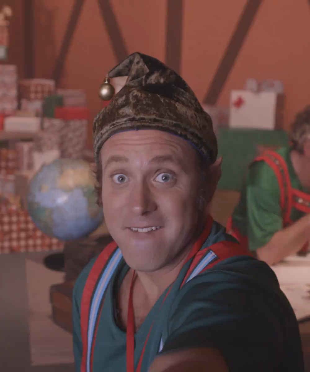 Man in a festive elf costume taking a selfie, wearing a velvet hat with a bell, with a globe and Christmas decorations in the background.