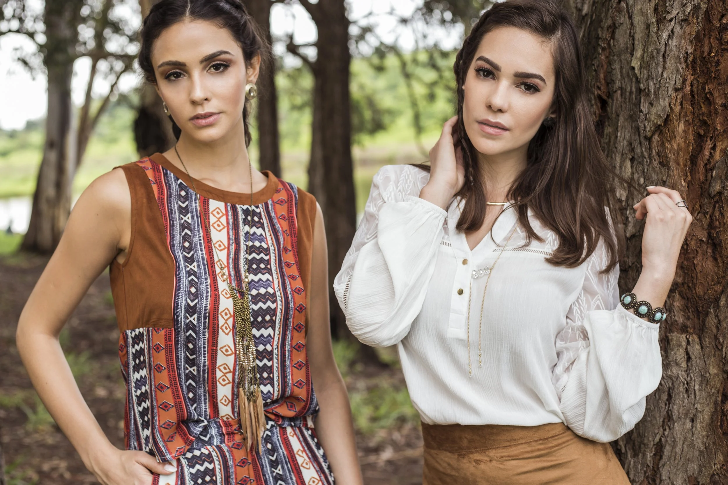 Two young women standing outdoors near trees, with a lake in the background. One wears a colorful patterned sleeveless dress, and the other wears a white blouse and brown skirt.
