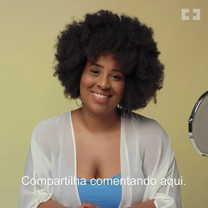 A smiling woman with curly hair wearing a white sheer cover-up and a blue top, standing in front of a mirror with a yellow background.