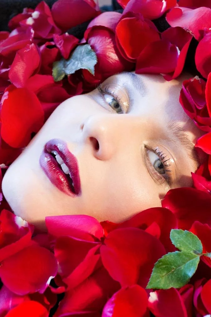 Close-up of a woman's face lying among red rose petals, with makeup and striking eyes.