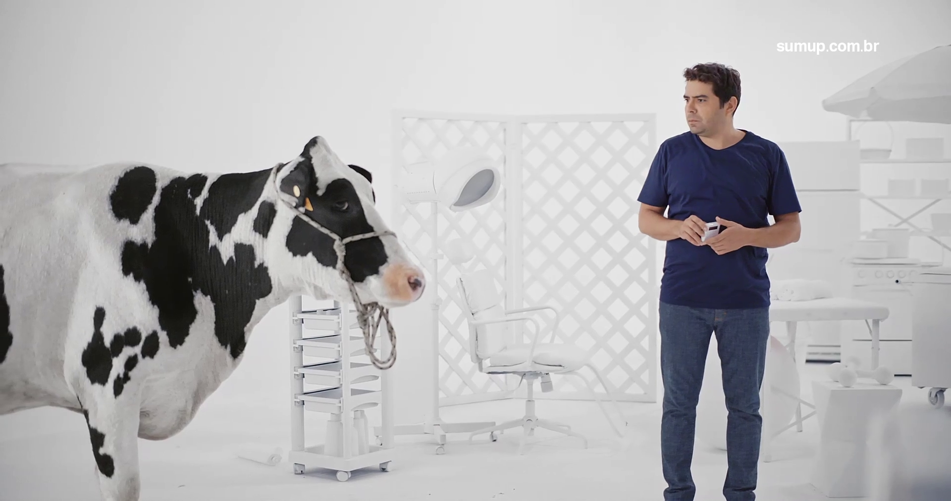 A man in a navy blue t-shirt and jeans holding a remote control stands next to a black and white cow in a bright white room with medical or laboratory equipment in the background.