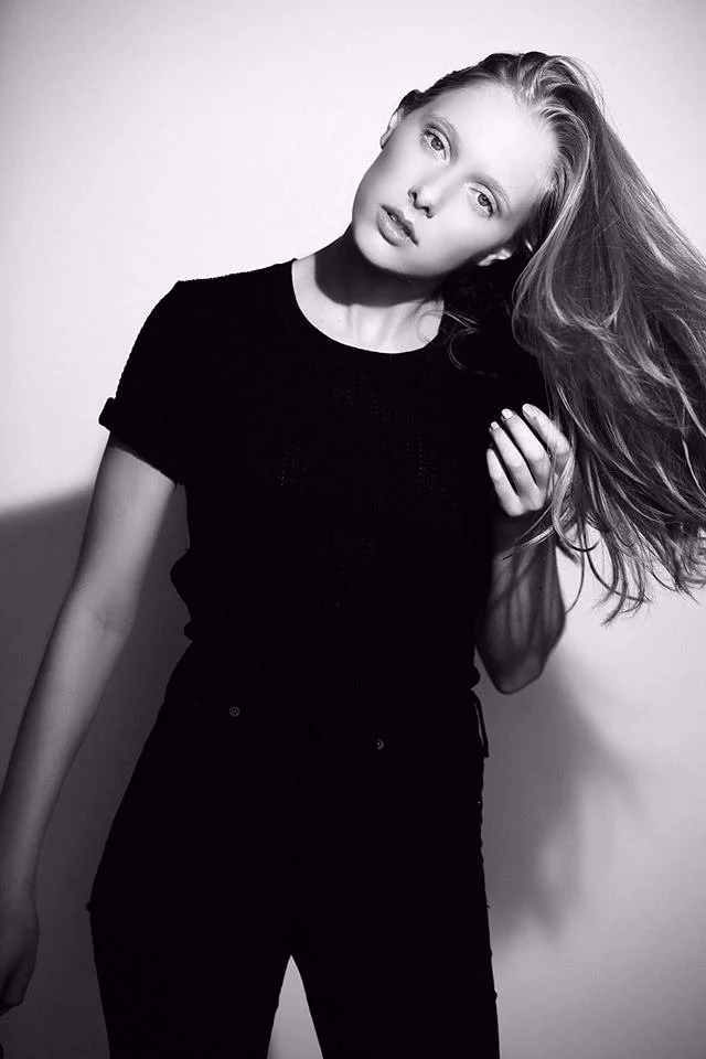 A young woman with long hair wearing a black t-shirt and pants, standing against a plain background, looking at the camera.