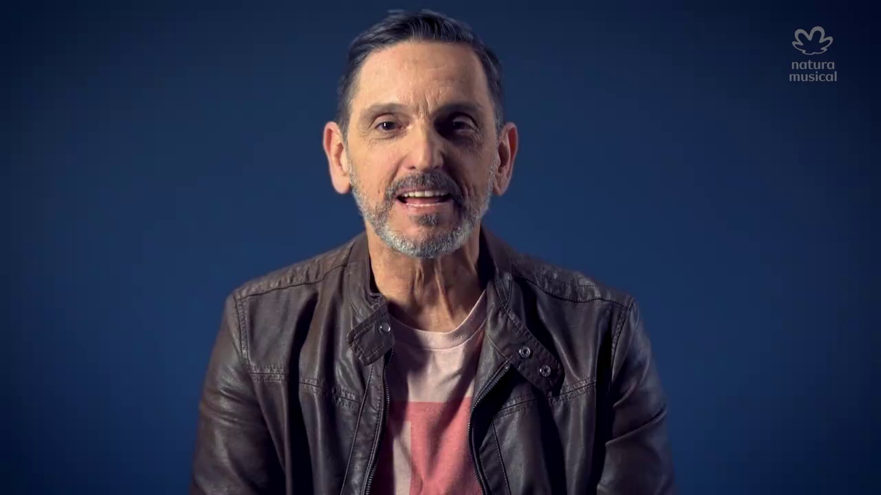 Close-up of a middle-aged man with short dark hair and a beard, wearing a brown leather jacket and a pink T-shirt, sitting against a dark blue background.