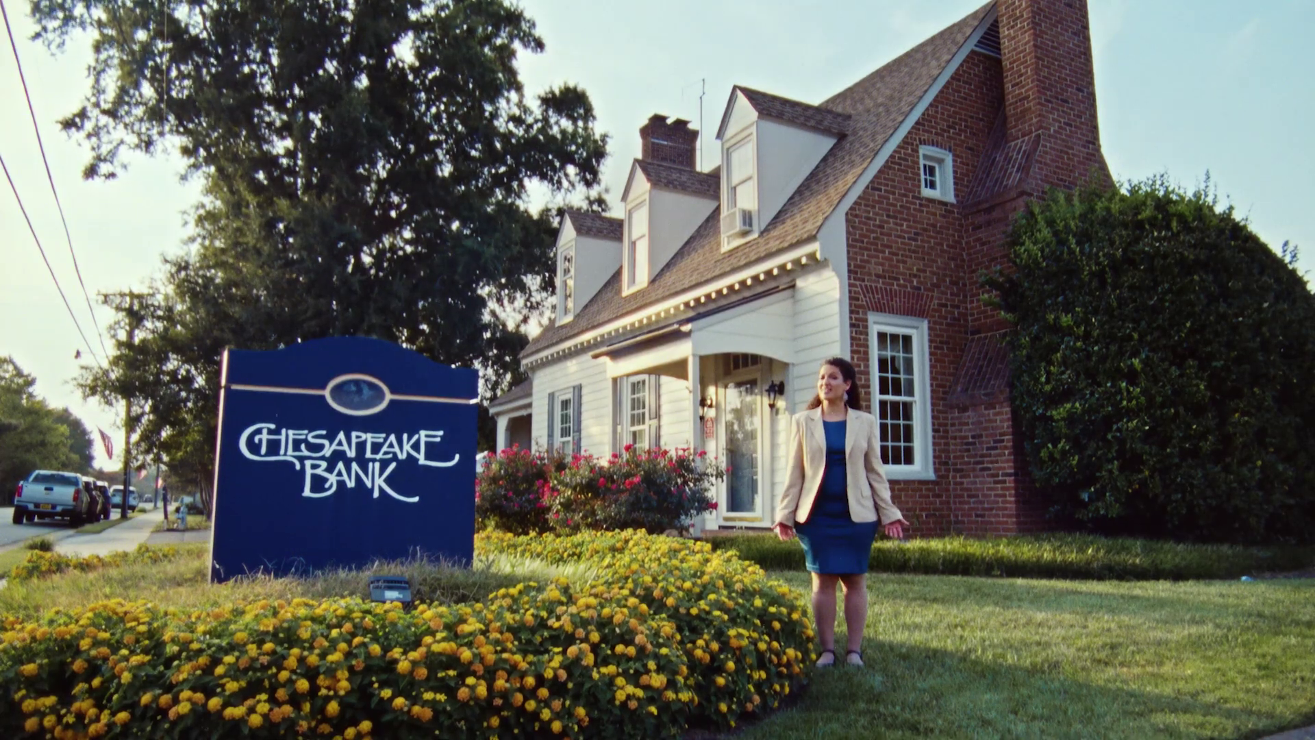 Woman in beige blazer and dark dress standing outside a white and red brick house with a Chester Pike Bank sign in front, surrounded by colorful flowers.
