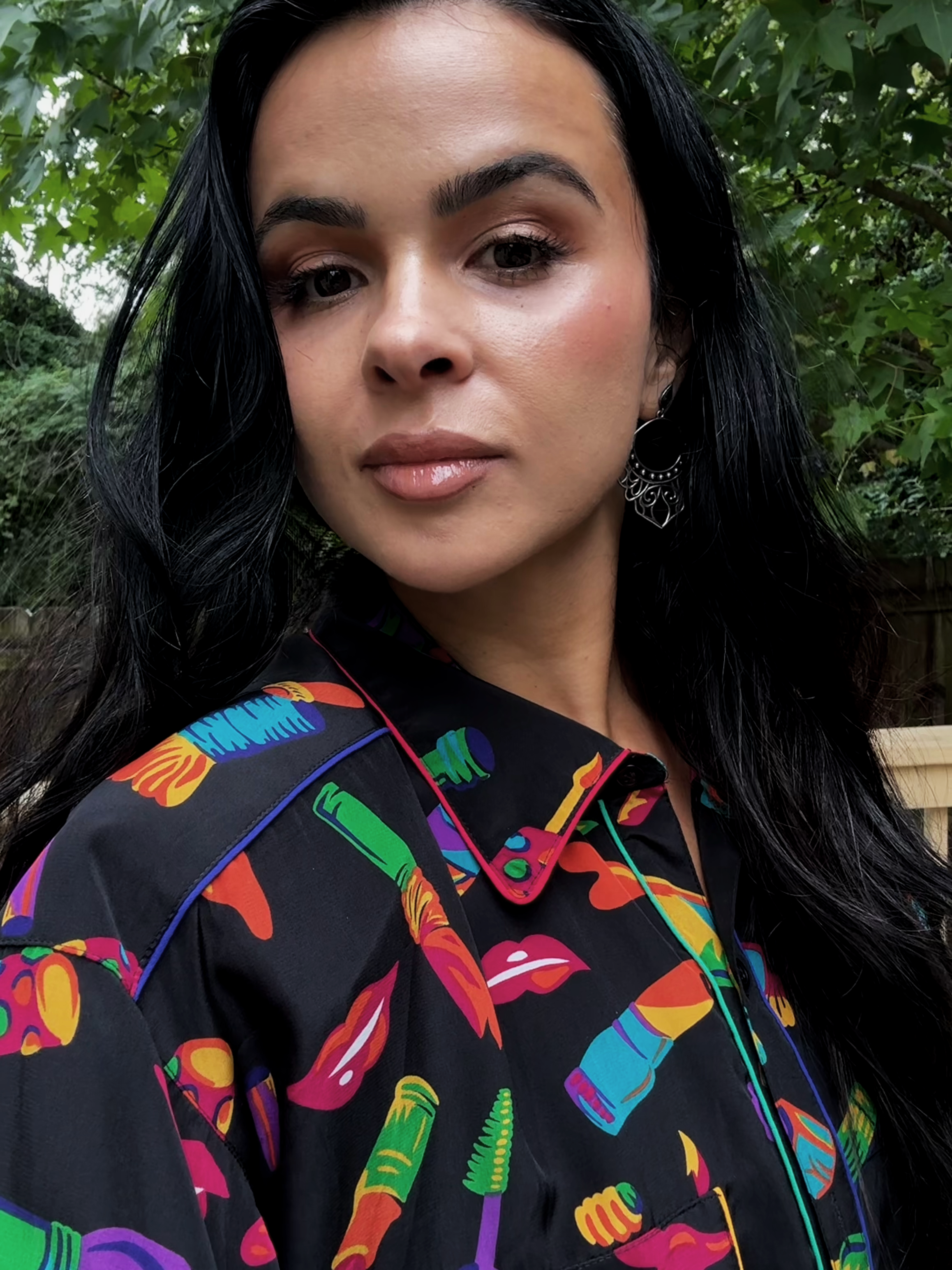 A woman with dark hair and earrings taking a selfie outdoors with green foliage in the background.