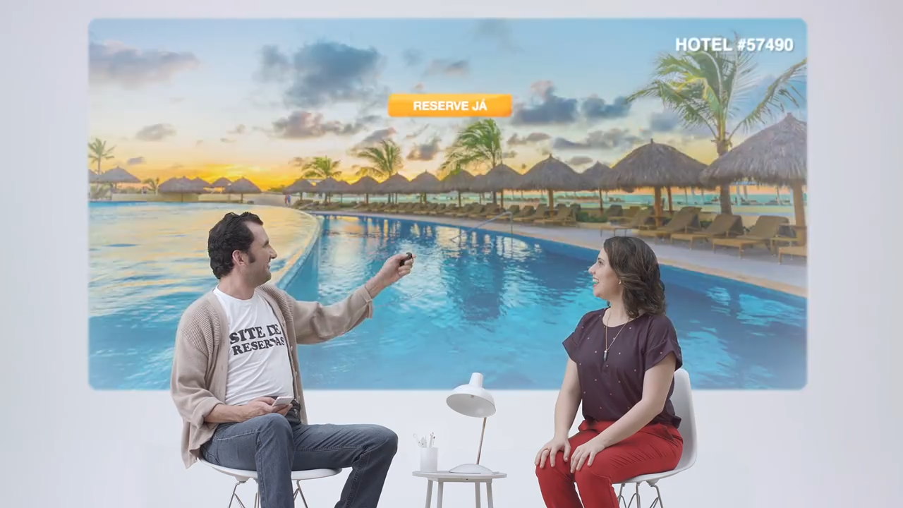 Two people sitting indoors, talking with a large poster of a tropical resort with a swimming pool, palm trees, and thatched huts in the background. The man is holding a small object and smiling, while the woman is listening. There is a small table wi