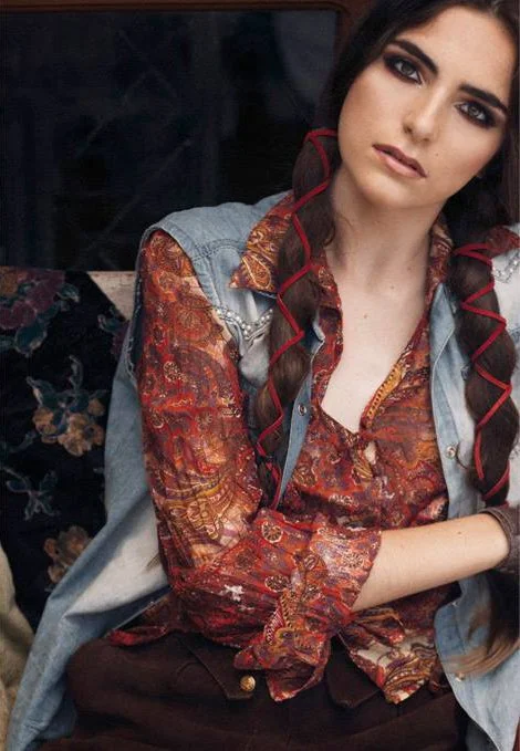 A young woman with dark hair, braided with red ribbons, and wearing a bohemian-style shirt with a paisley pattern, sitting on a vintage floral sofa.