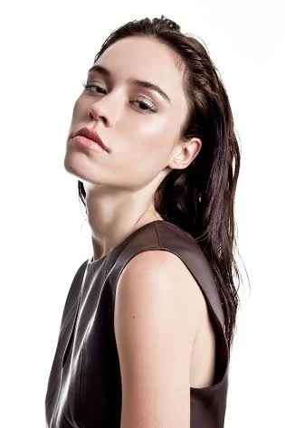 A young woman with fair skin and long dark hair posing with a serious expression, wearing a sleeveless top.