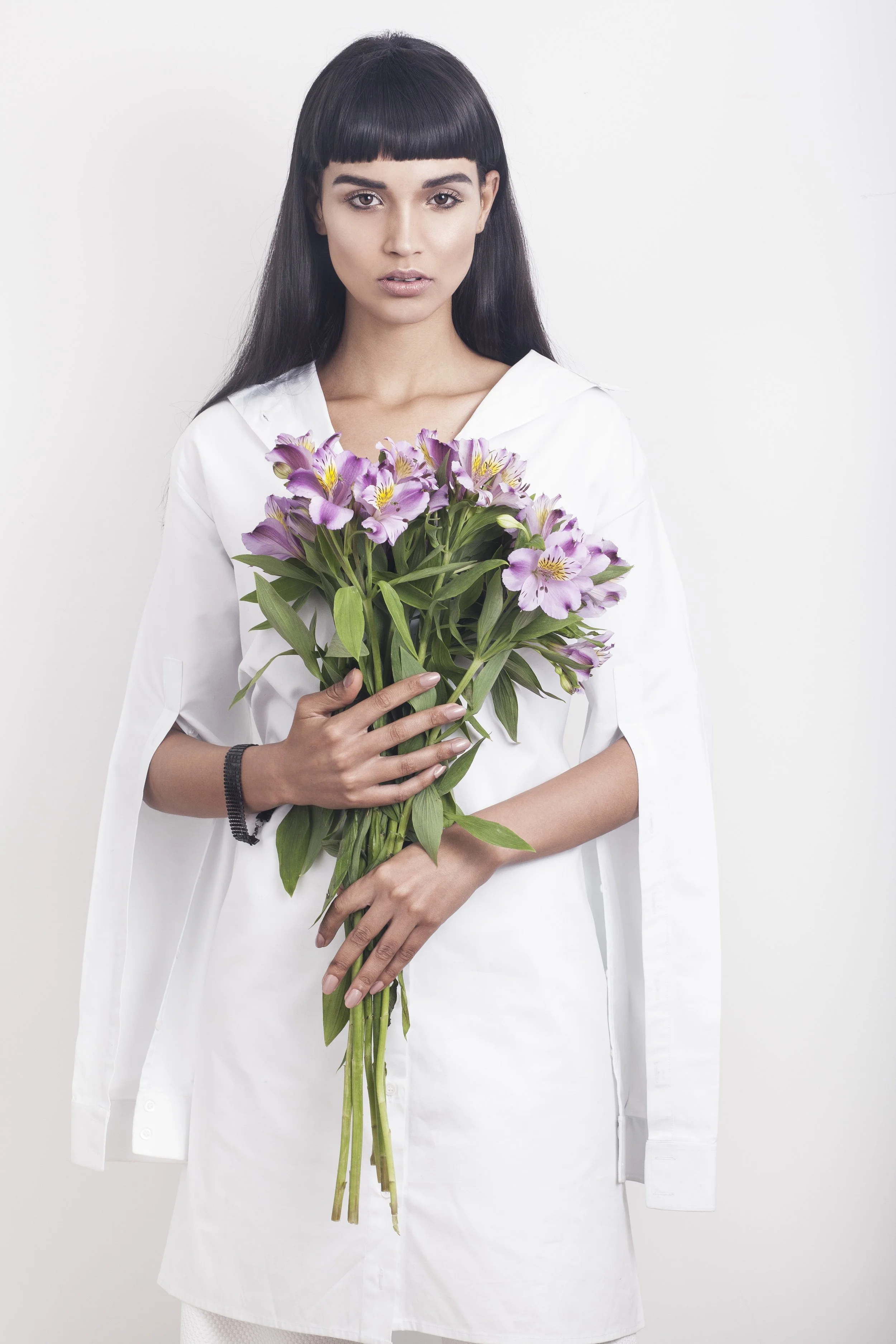 A woman with long dark hair and bangs holds a large bouquet of purple flowers while looking at the camera, against a plain white background.